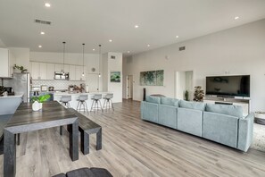 Eine moderne Küche mit weißen Schränken, ein Essbereich mit einem Holztisch und Bänken, ein graues Sofa und ein Flachbildfernseher.