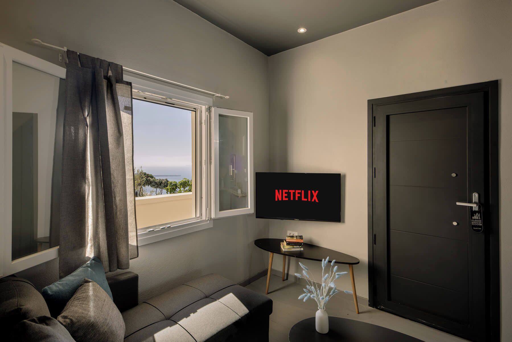 Ein modernes Wohnzimmer mit einem Fernseher, der 'NETFLIX' zeigt, einem Sofa, einem kleinen Tisch mit Büchern und einer Tür.