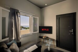 Ein modernes Wohnzimmer mit einem Fernseher, der 'NETFLIX' zeigt, einem Sofa, einem kleinen Tisch mit Büchern und einer Tür.