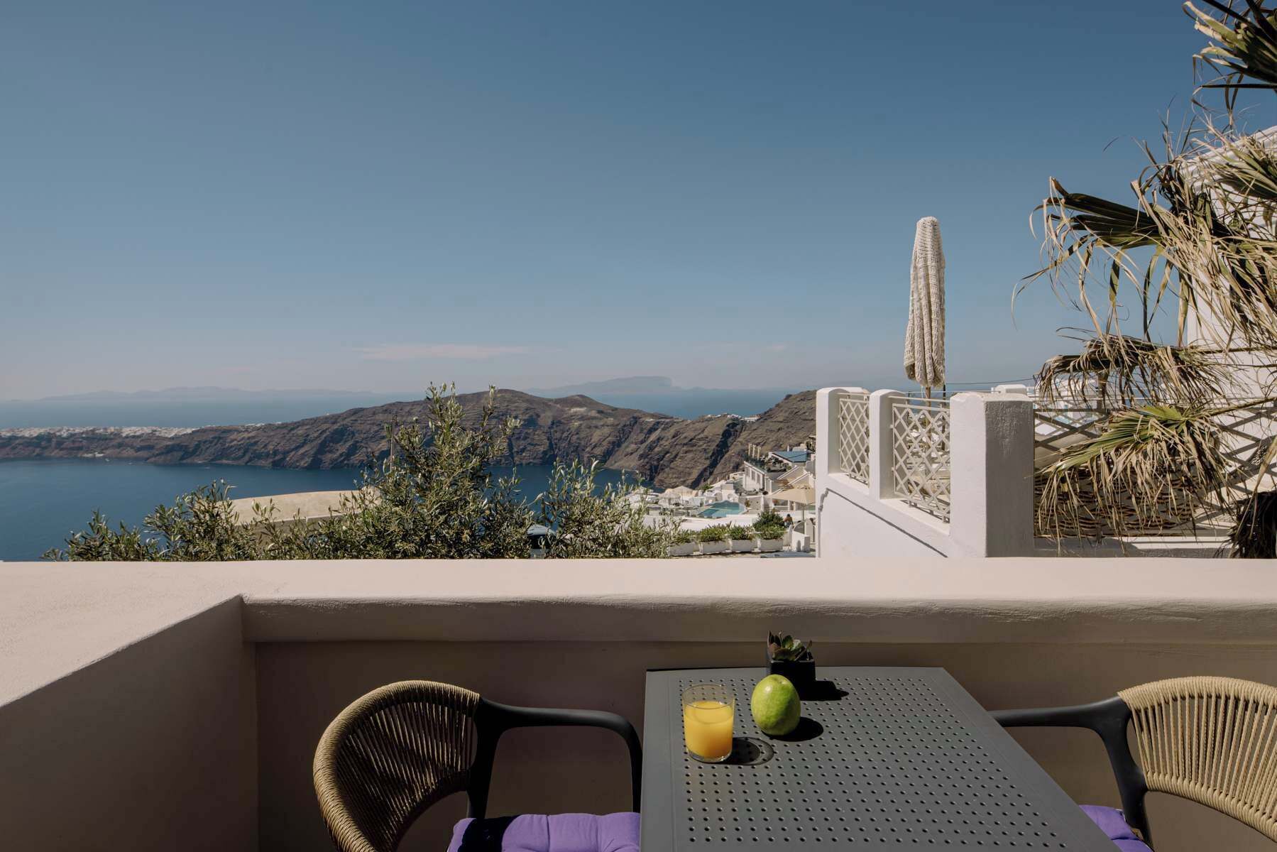Un balcon avec vue sur la mer et les collines, une table avec un verre de jus d’orange et un fruit, et des chaises en osier.