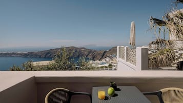 Un balcon avec vue sur la mer et les collines, une table avec un verre de jus d’orange et un fruit, et des chaises en osier.