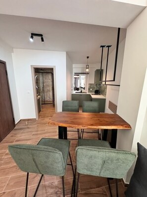 Ein moderner Essbereich mit einem Holztisch und vier grünen Stühlen, im Hintergrund eine Küche und ein Flur, der zu einem Badezimmer führt.