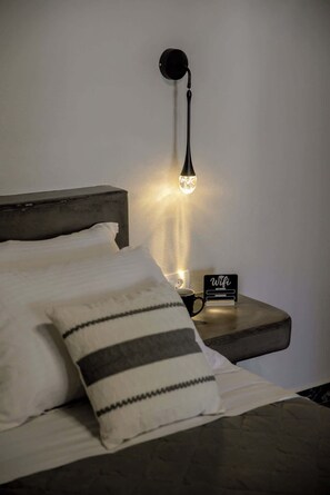 A bed with pillows, a bedside table with a lamp, a sign, and a cup. - Grande Studio with Jacuzzi Nea Premium Residences (Imerovigli)