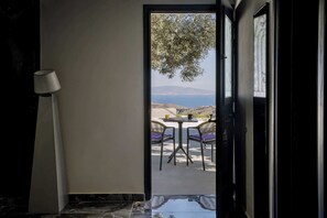 An open doorway leading to a terrace with a table and chairs, a lamp, and a view of the sea and hills. - Grande Studio with Jacuzzi Nea Premium Residences (Imerovigli)