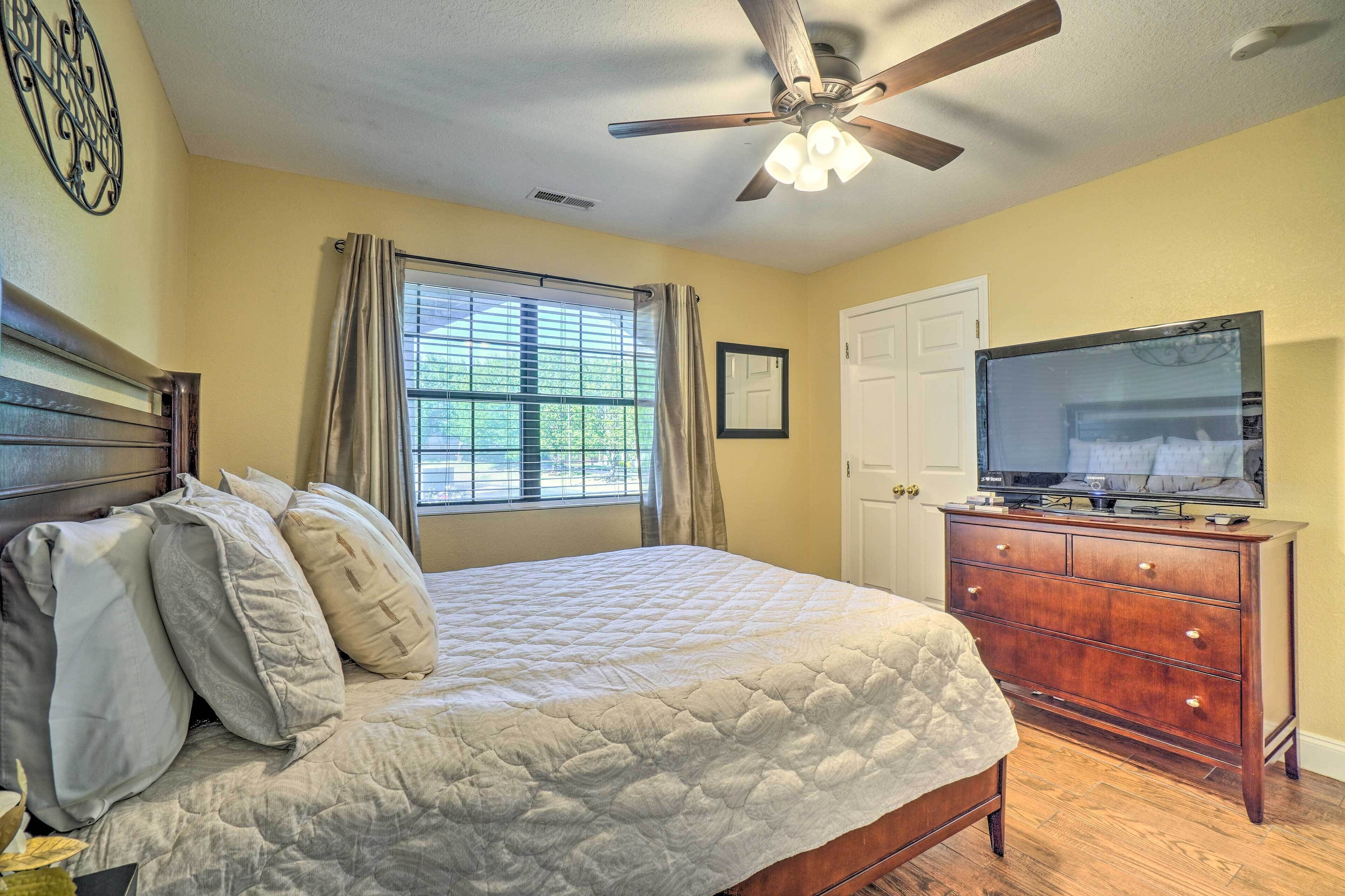 A bedroom with a bed, a dresser, a ceiling fan, a window with curtains, and a television.