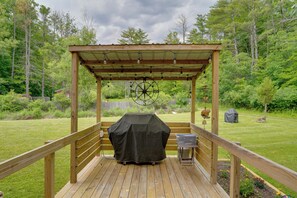 Eine Holzterrasse mit überdachter Grillgelegenheit, einer Pergola mit Lichterketten und Blick auf einen grünen Garten mit Bäumen.