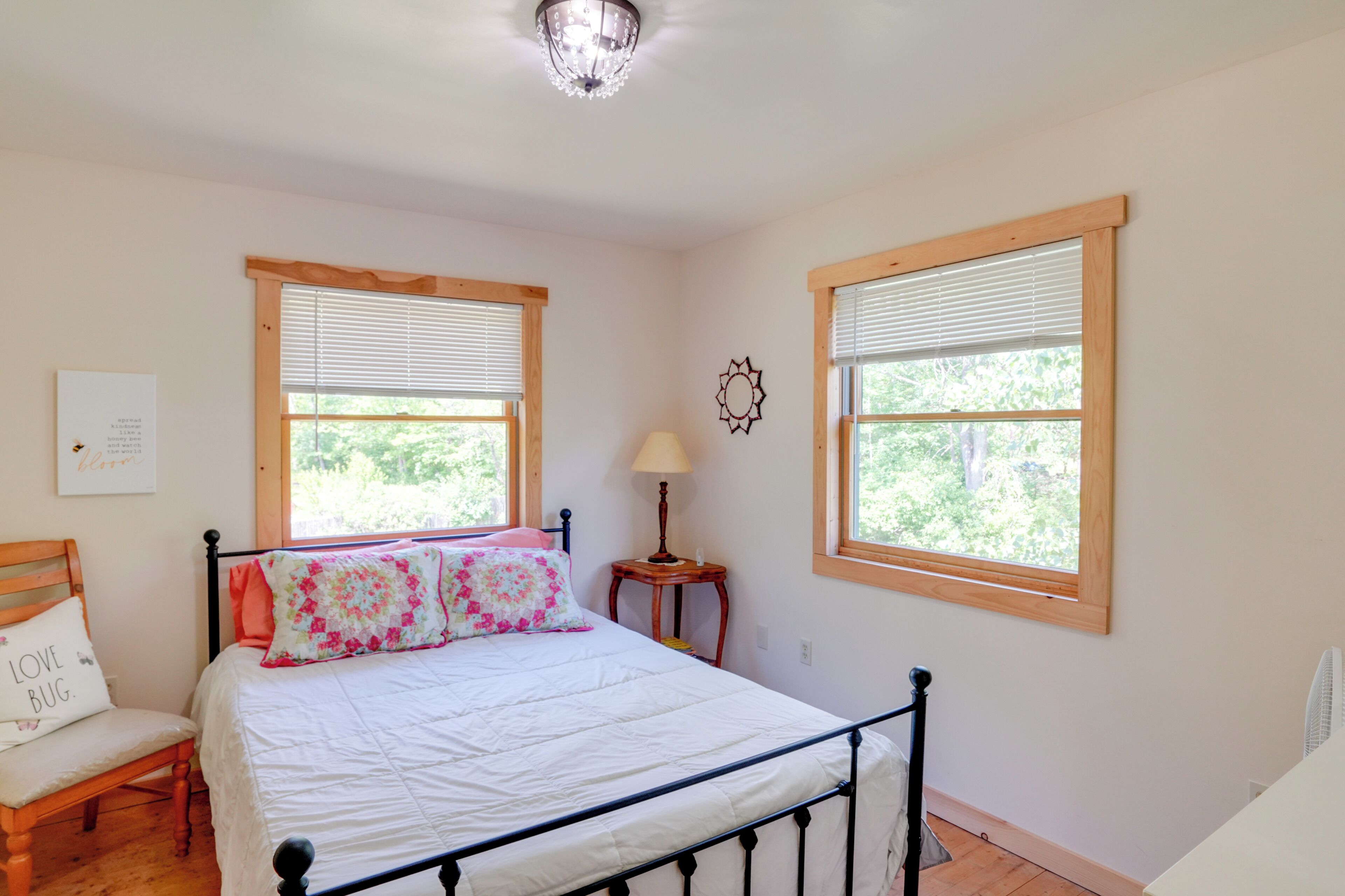 A bedroom with a bed, a chair, a nightstand, a lamp, and two windows with blinds.