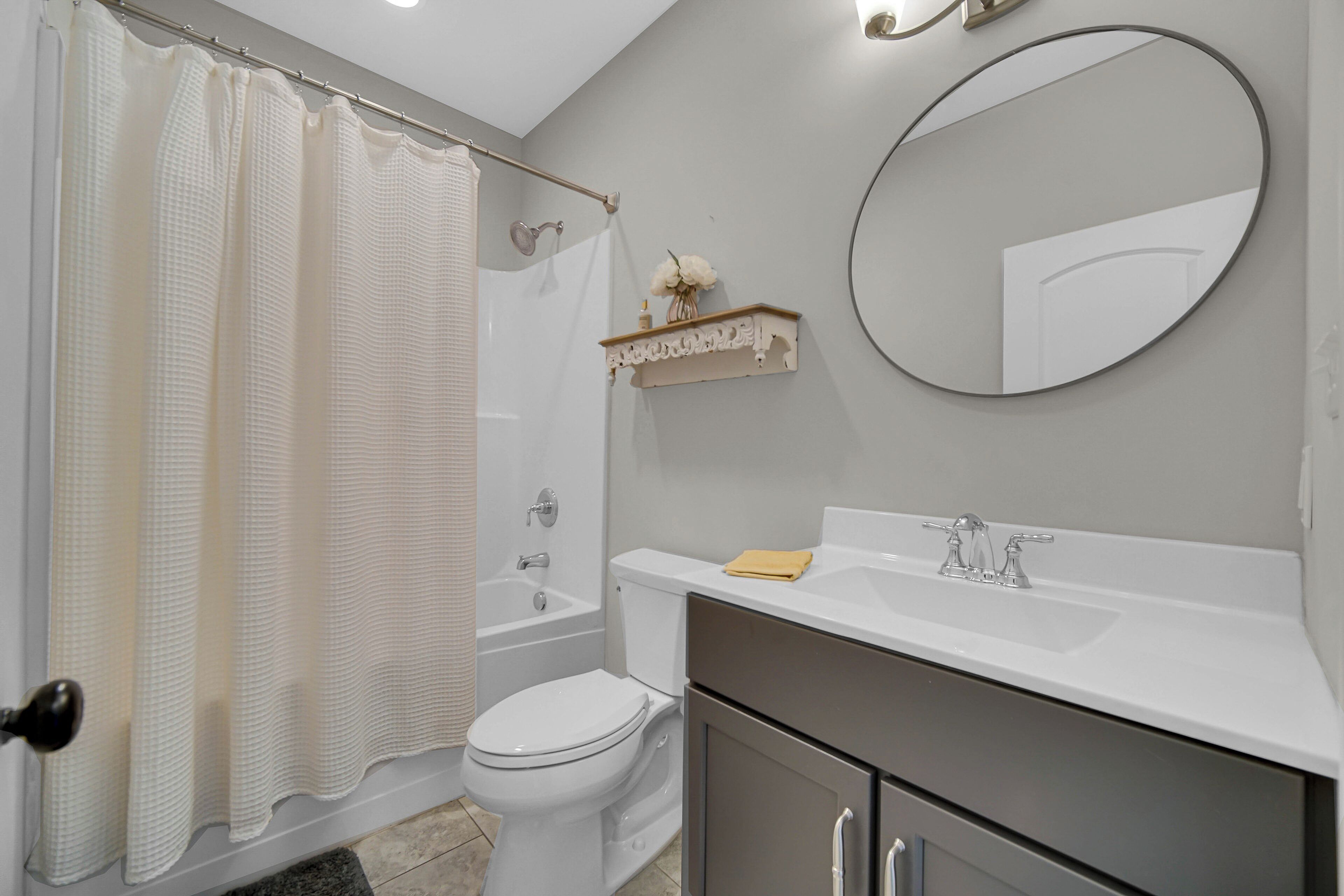 A bathroom with a shower curtain, toilet, bathtub, sink, mirror, and a shelf with a vase.