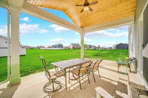 Une terrasse couverte avec une table et des chaises, un ventilateur de plafond et une vue sur une pelouse verdoyante et des maisons.