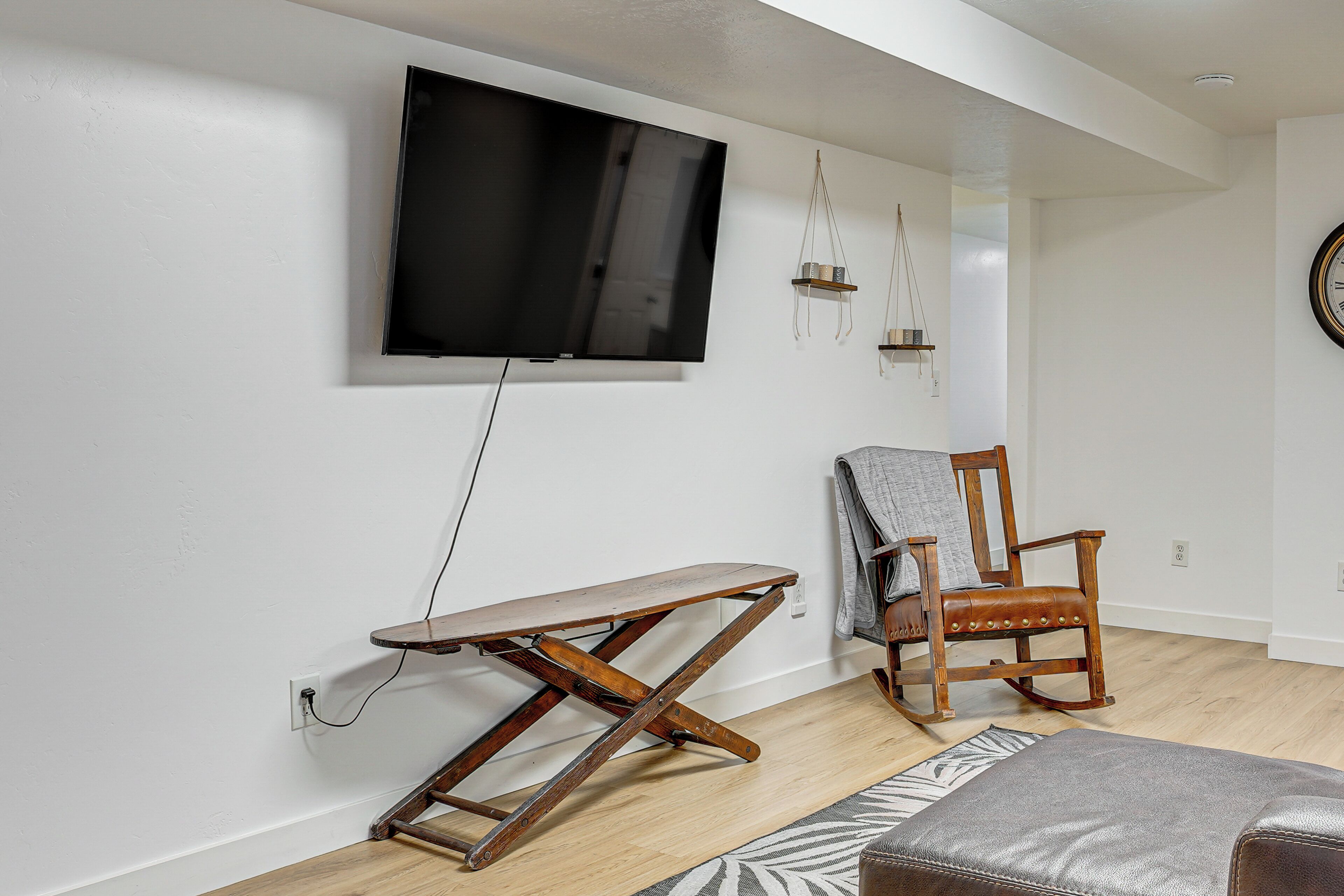Una sala de estar con un televisor montado en la pared, un banco de madera, un sillón mecedora y una alfombra estampada.