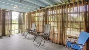 Un espacio exterior techado con sillas de metal y un banco, ventanas amplias con cortinas y piso de concreto.
