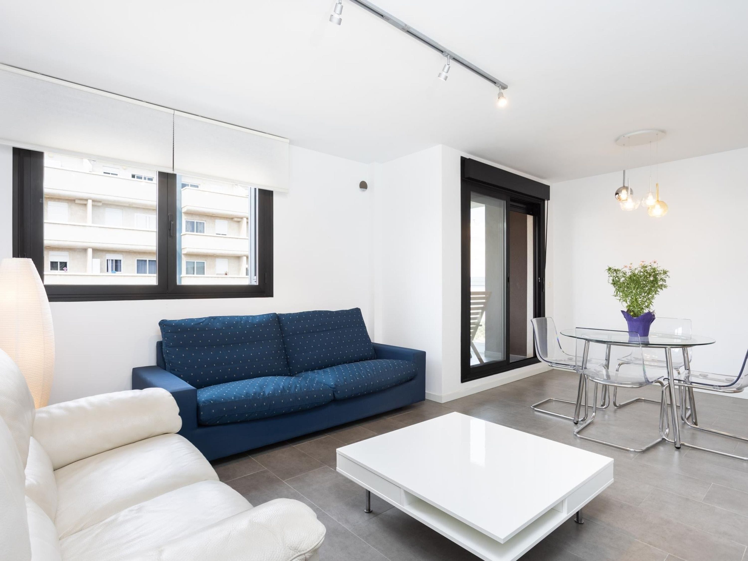 A modern living room with a sofa, a coffee table, a dining table with chairs, a potted plant, and a view of the outside.