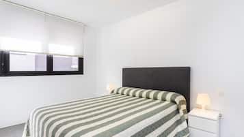 Een slaapkamer met een bed, nachtkastje, een aan de muur bevestigde televisie en een raam met jaloezieën.