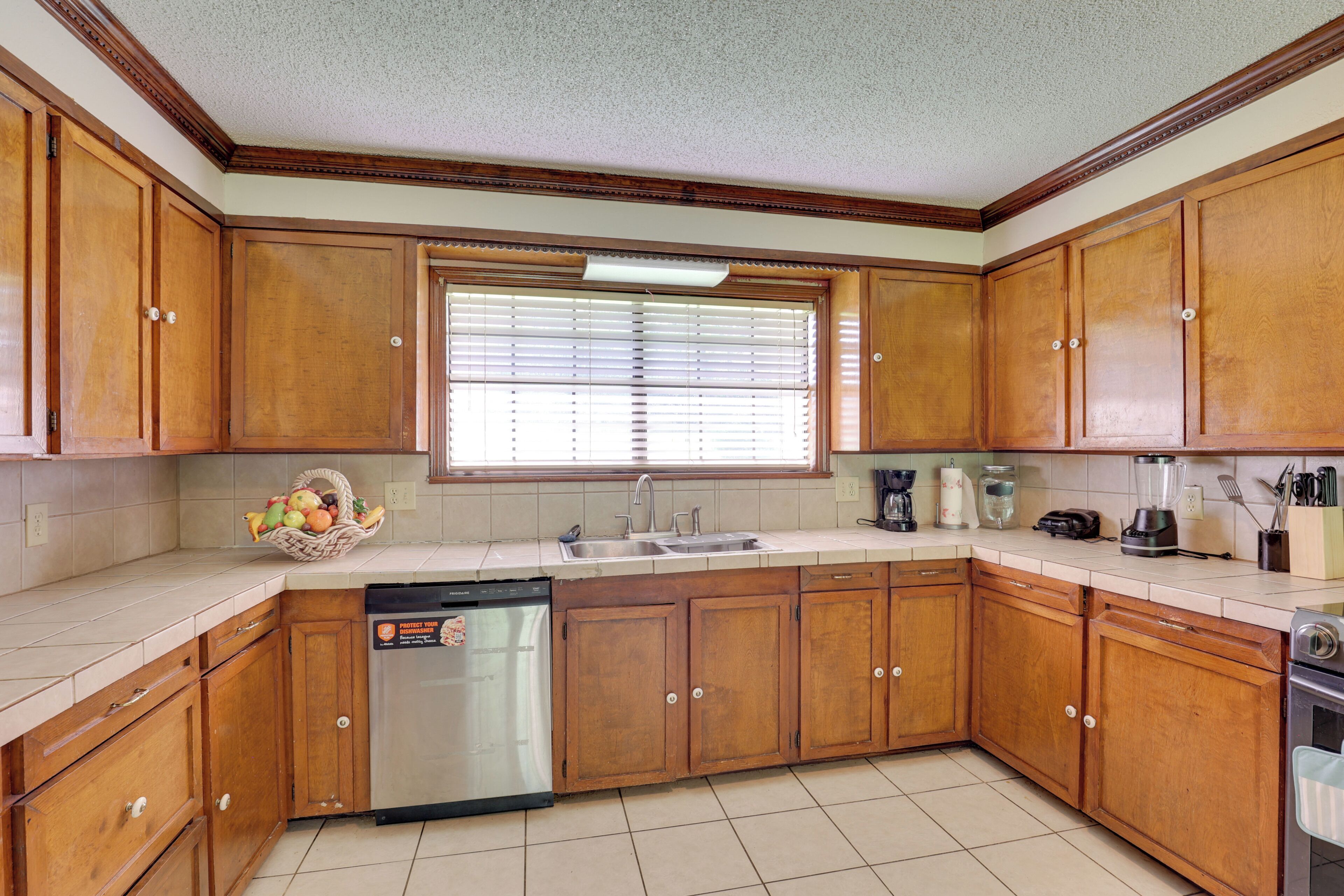 A kitchen with wooden cabinets, a dishwasher, a window with blinds, and a countertop with various appliances and utensils.