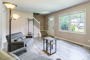Ein Wohnzimmer mit grauem Ecksofa, einem Metall-Couchtisch, einer Treppe und einem großen Fenster mit Blick auf Grünflächen.