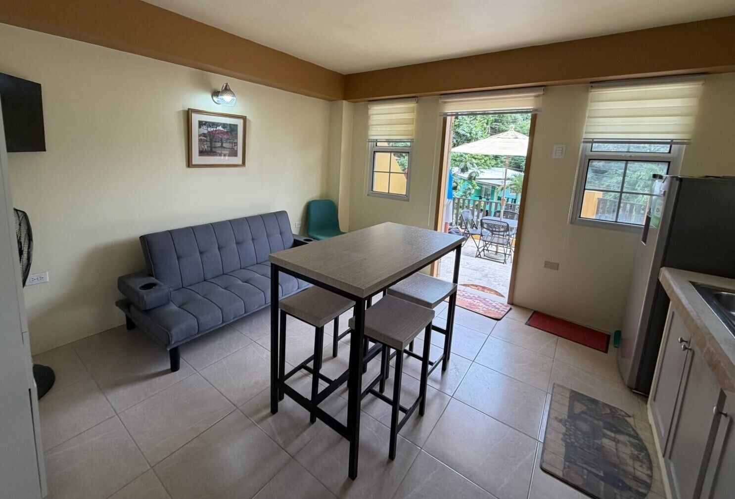 Un espace de vie compact comprenant un canapé, une table à manger avec des chaises, un coin cuisine et une porte-fenêtre coulissante donnant sur un espace extérieur.