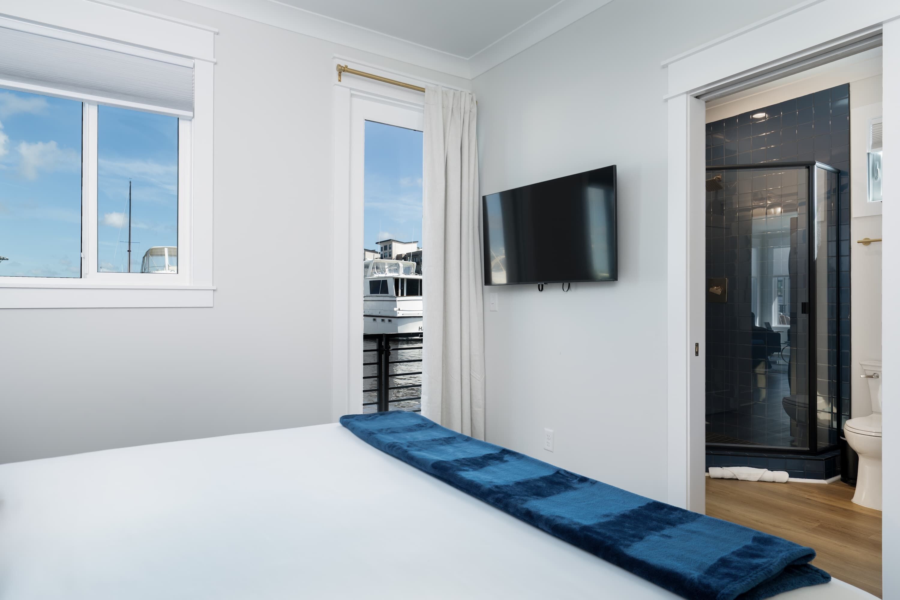 A bedroom with a bed, a TV mounted on the wall, and a view of boats through a window.