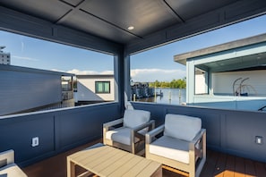 Ein überdachter Außenbereich mit zwei weißen Stühlen und einem Holztisch, mit Blick auf einen Pool und ein Gebäude mit weißer Fassade.