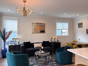 A modern living room with a black sofa, teal armchairs, a glass coffee table, and a chandelier.