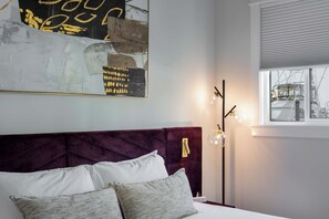 Ein Schlafzimmer mit einem Bett, Kissen, einem Wandbild, einer Stehlampe und einem Fenster mit Jalousien.