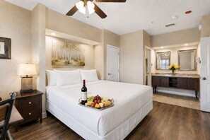 Ein Hotelzimmer mit Bett, Nachttisch, Lampe, Gemälde, Esstisch mit Früchten und Wein, Badezimmer mit Spiegel und Deckenventilator.