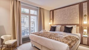 Quarto de hotel com uma cama grande, uma cadeira, uma janela com cortinas e um ponto de luz fixado na parede.