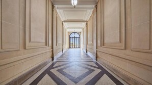 Um corredor com piso geométrico, ladeado por paredes bege com molduras decorativas, conduzindo a uma porta de vidro.