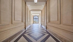 A corridor with a geometric patterned floor, flanked by beige walls with decorative moldings, leading to a doorway with a glass door. - THE ESSENCE OF PARISIAN ELEGANCE ,JUST MOMENTS FROM THE ICONIC ARC DE TRIOMPHE (Paris)