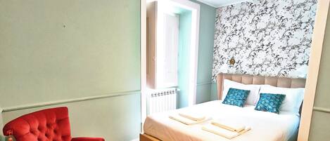 Un dormitorio con una cama, una silla roja, una mesita con un florero y una pared con estampado floral.