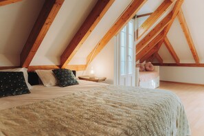 Un dormitorio con techo inclinado, vigas de madera, una cama con ropa de cama estampada, una mesita de noche con una lámpara y una ventana.