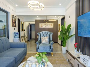 A modern living room with a blue sofa, a tufted armchair, a coffee table with decorative items, and a large potted plant. - 2-bedroom apartment in lavington Nairobi. With swimming pool and gym. Near malls (Nairobi)