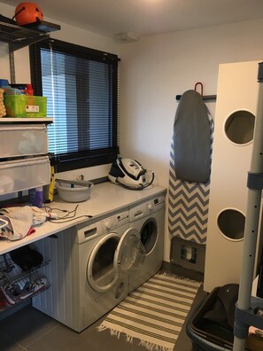 A laundry area with a washing machine, dryer, ironing board, and a window with blinds. - Quiet Villa with Pool - Saint Nolff (Saint-Nolff)