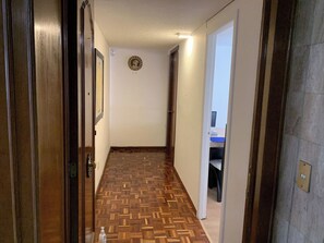 Ein Flur mit einem gemusterten Holzboden, einer Tür links und einem Schreibtisch mit Stuhl im Hintergrund.