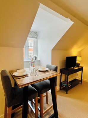 A dining area with a table set for four, a television on a stand, and a window with a view of a building.