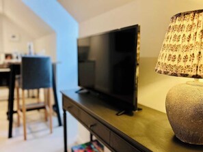 Ein modernes Wohnzimmer mit einem Flachbildfernseher auf einem Holztisch, einer gemusterten Lampe und einem Essbereich im Hintergrund.