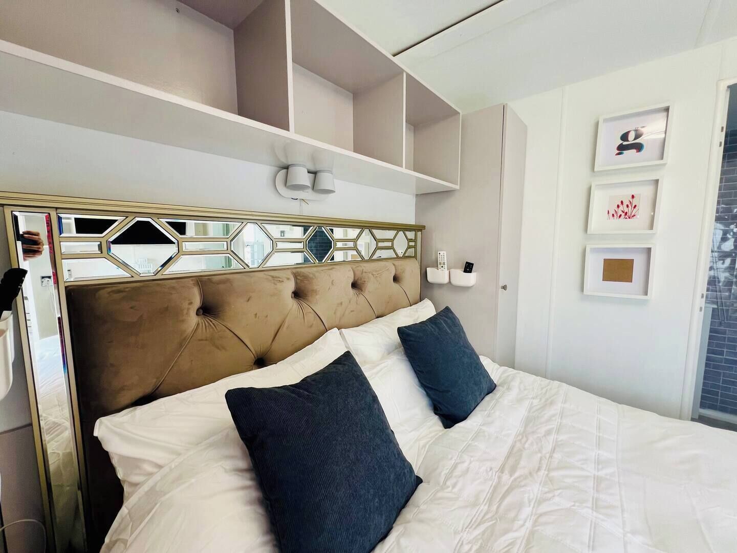 Ein Bett mit gestepptem Kopfteil, weißer Bettwäsche und zwei dunklen Kissen. An der Wand befinden sich Regale mit gerahmten Bildern sowie eine Wandleuchte.