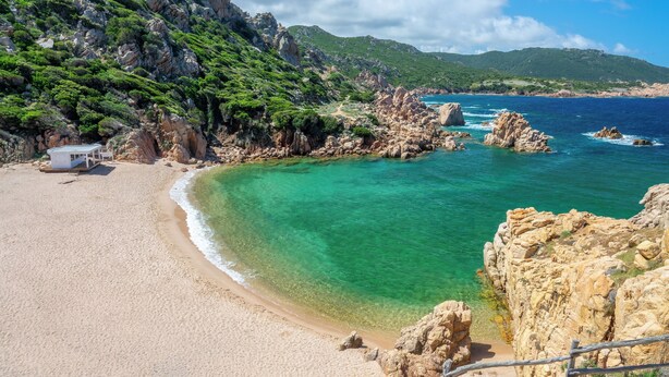 Una spiaggia con acque turchesi cristalline, scogliere rocciose e un piccolo edificio sulla sabbia.