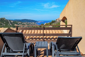 Eine Terrasse mit zwei Liegestühlen und einem Tisch, mit Blick auf eine Küstenlandschaft mit Gebäuden und Hügeln.