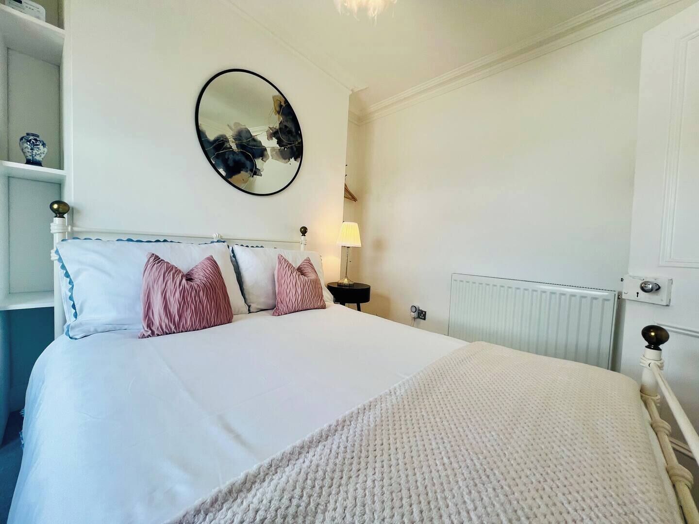 A neatly made bed with a white duvet, a round mirror on the wall, a bedside lamp, and a radiator.