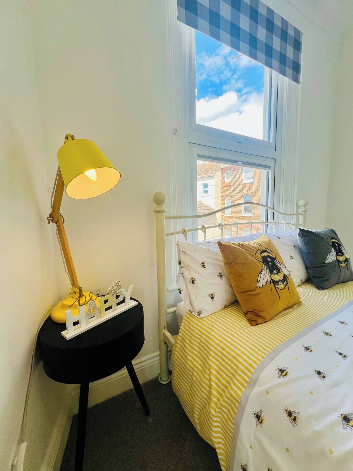 A bedroom with a bed, a bedside lamp, a window with a view of buildings, and a small table with a lamp.