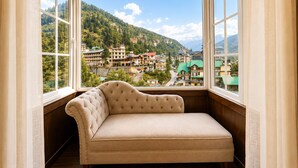 A room with a window view of a mountainous town, a cushioned bench, and a hanging light fixture. - The himalayan chester (Manali)