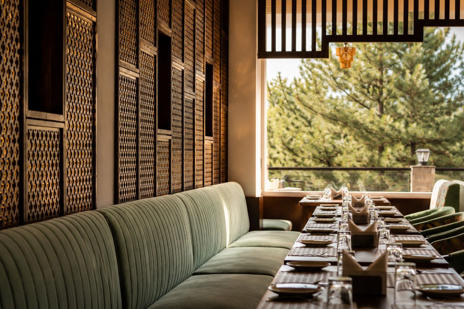 A dining area with a long table set for a meal, a green upholstered bench, and a view of trees through a window.