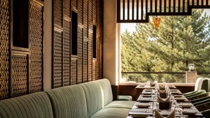 A dining area with a long table set for a meal, a green upholstered bench, and a view of trees through a window. - The himalayan chester (Manali)