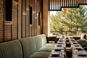 Un comedor con una mesa larga dispuesta para una comida, un banco tapizado en verde y vistas a árboles a través de una ventana.