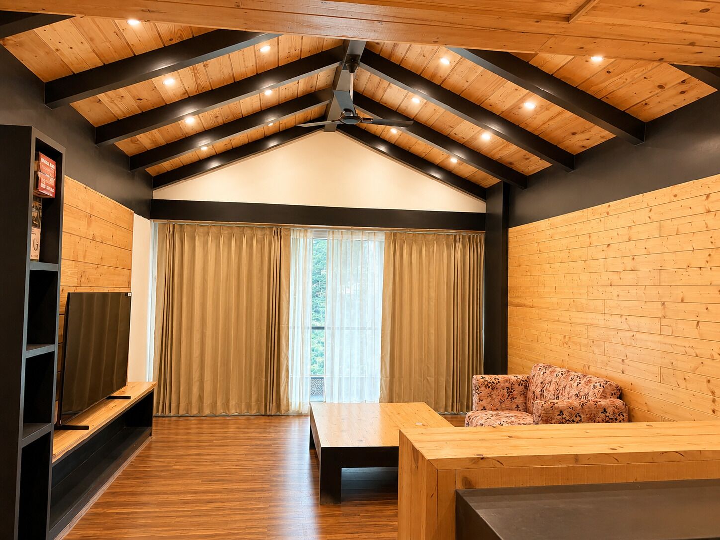 Una habitación con techo inclinado, vigas de madera, un sofá, una mesa y un amplio ventanal con cortinas.