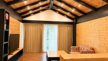 Una habitación con techo inclinado, vigas de madera, un sofá, una mesa y un amplio ventanal con cortinas.