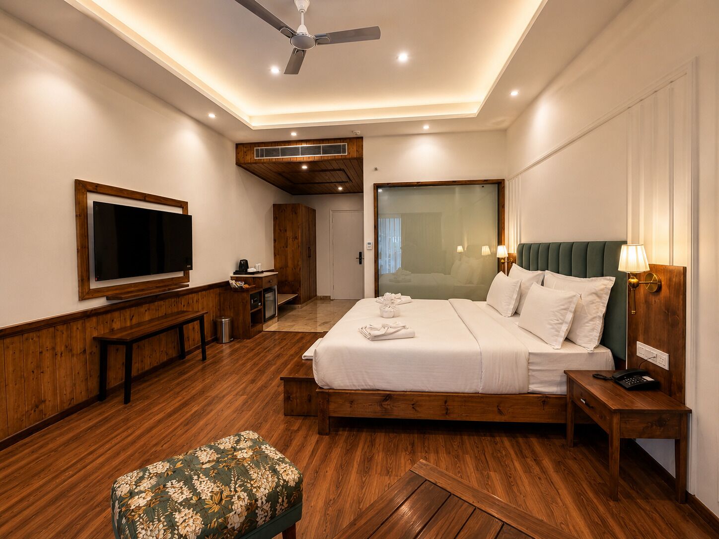 A hotel room with a bed, a TV, a desk, a chair, a bench, and a ceiling fan.