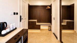 A hotel room with a wooden wardrobe, a small seating area, a coffee station, and a trash bin.
