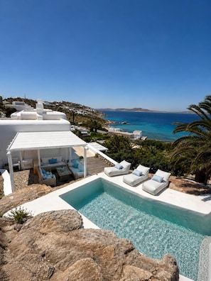 Ein Poolbereich mit Liegestühlen, einer weißen Pergola und Blick auf Meer und Hügel.