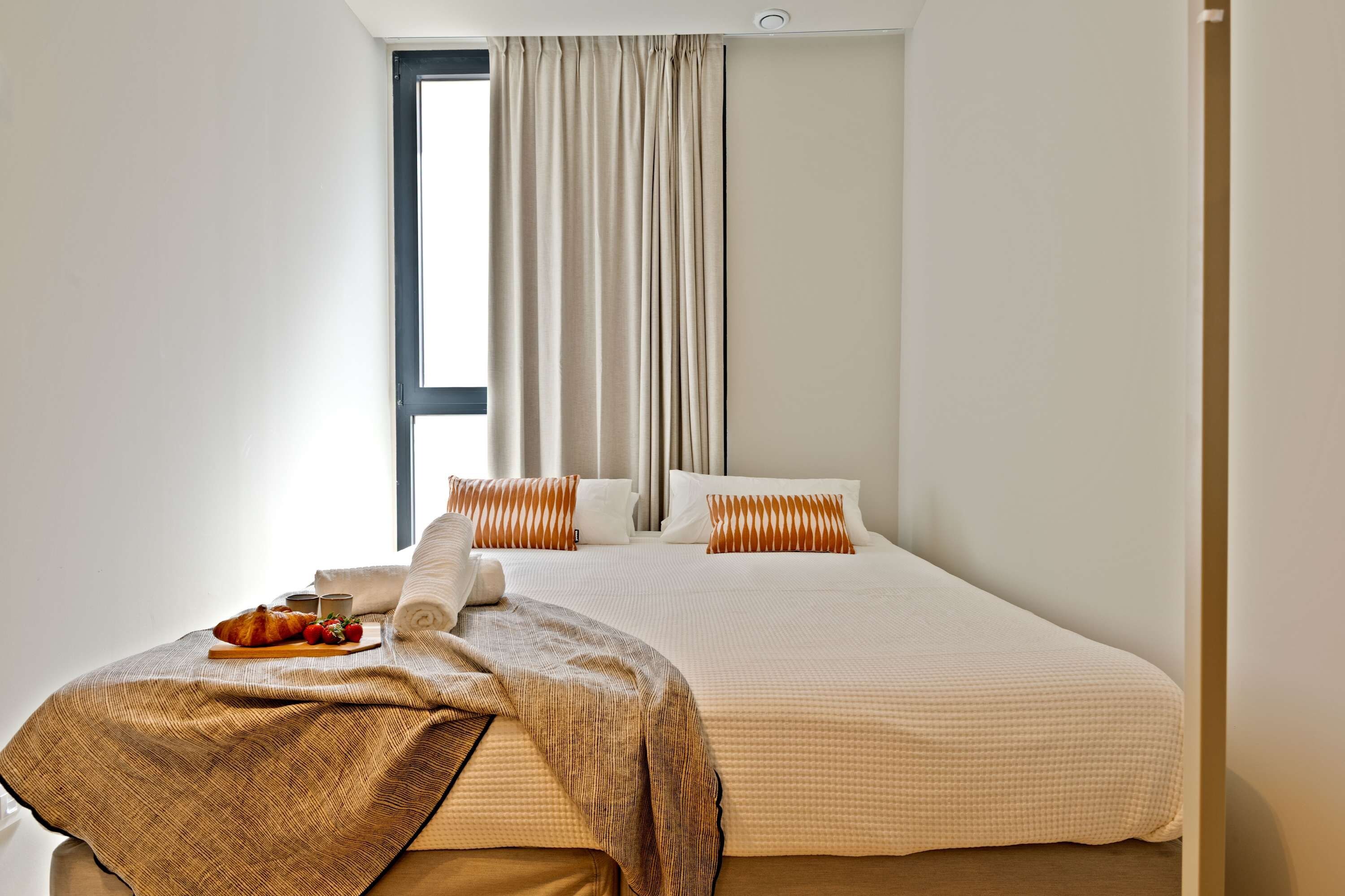 A hotel room with a bed, a window with curtains, a bedside table with a tray of food, and a blanket.